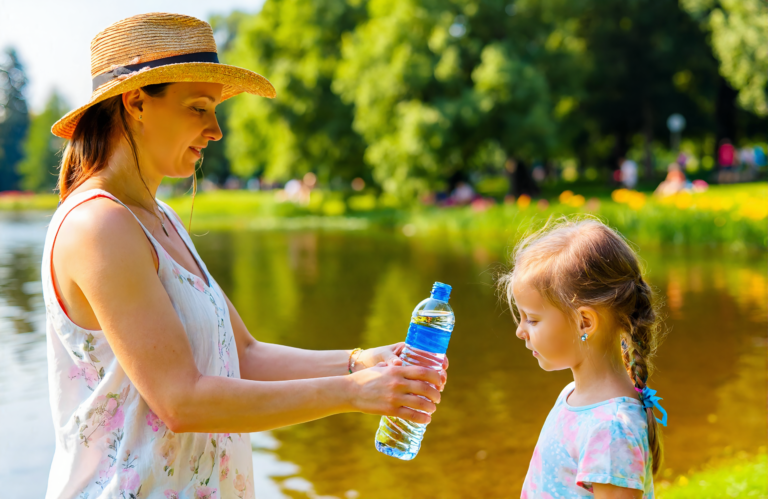 White mother in a straw sun hat handing a water bottle to her young daughter at a Northshore lakefront park in summer heat