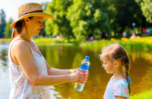 White mother in a straw sun hat handing a water bottle to her young daughter at a Northshore lakefront park in summer heat