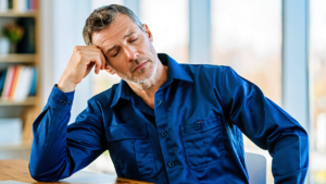 Tired man in a navy work shirt pressing a hand to his temple at a home office desk, the kind of fatigue that leads people to look up B12 shots near me