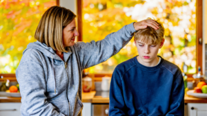 Mother in a gray zip-up hoodie resting her hand on her pale, tired teenage son's head at a kitchen counter on a fall morning