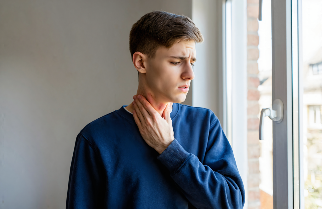 Young man in a navy sweatshirt pressing his fingers to the side of his neck by a window, checking for swollen glands, a common sign of mono