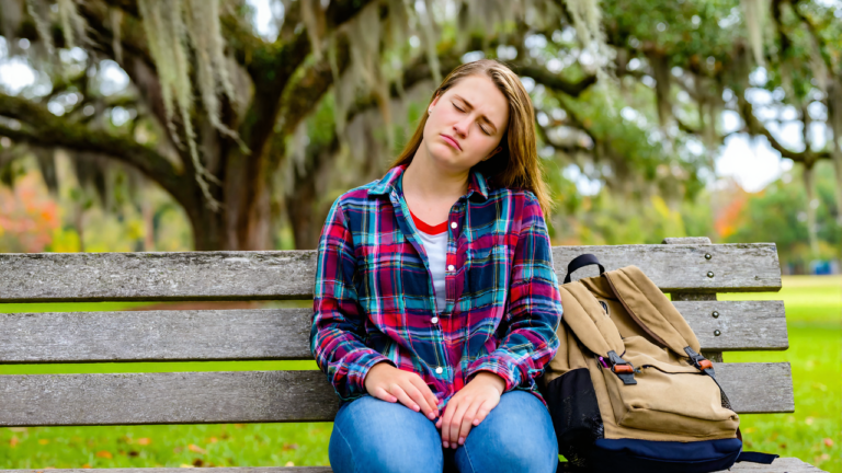 Tired young woman in a plaid flannel shirt resting on a park bench under live oaks, showing the fatigue that is common with mono symptoms