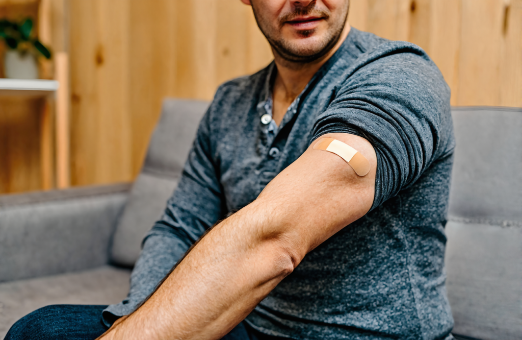 Man in a gray henley sitting on a sofa with his sleeve rolled up and a small bandage on his upper arm after an STD testing visit