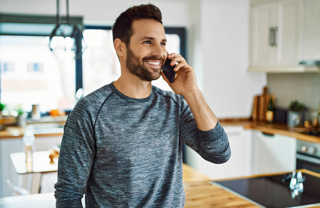 Man in a gray long-sleeve shirt smiling on a phone call in a bright kitchen while planning TB test visits near Covington, LA
